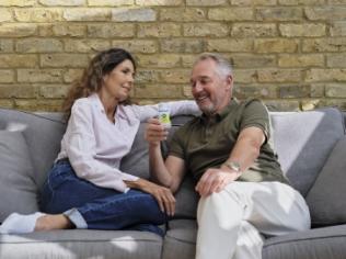 An elderly couple relaxing on a grey sofa. The man is holding a small glass of yellow Souvenaid, about to take a drink whilst smiling.